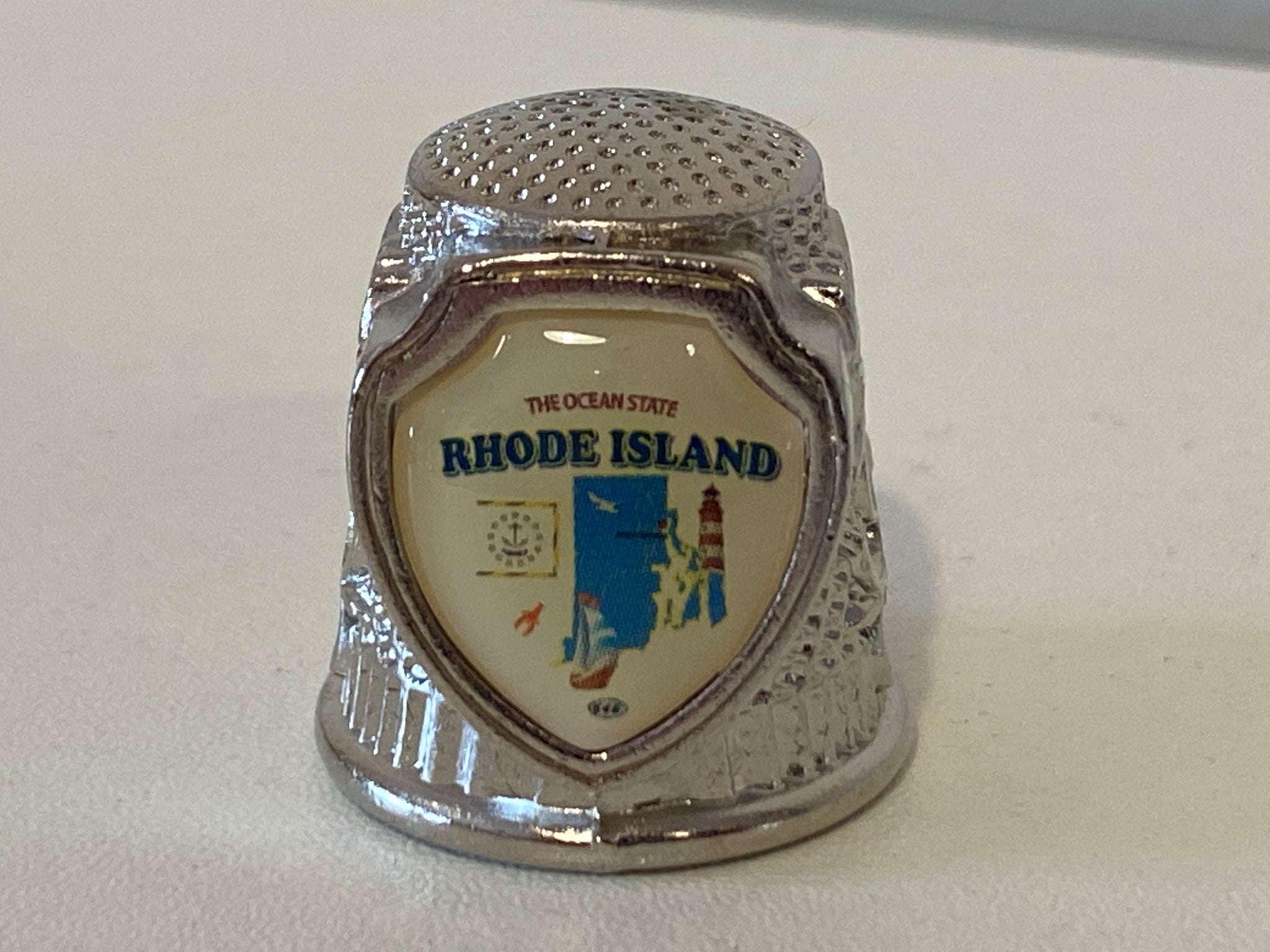 This ceramic thimble features the state of Rhode Island along with a red hen and some flowers. Gold line around the bottom and typical thimble bumps on the top. This is an adorable little addition to your sewing room, or thimble collection!